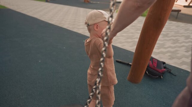 Young child in brown outfit and cap holds hands with adult near playground equipment including swings. scene takes place outdoors on soft ground surface with background elements like benches