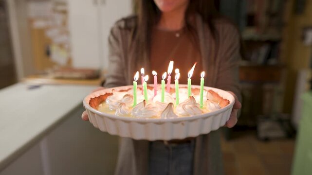 A mother walks through a modern rustic kitchen carrying a lemon pie cake with lit candles, singing happy birthday as she heads off-frame. Camera opens from close-up. Slow motion, video 4K