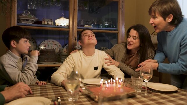 A teenage boy closes his eyes making a wish while his cheering family applauds and hugs him before he blows out the tiramisu cake candles. Slow motion, video 4K.
