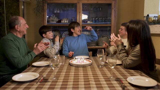 Grandchildren and family gather around grandmother, applauding and singing as she makes a wish and blows out the candles on her lemon pie birthday cake. Camera pushes in slowly. Video 4K.