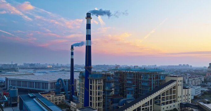 Aerial view of an industrial manufacturing plant with tall blue and white smokestacks emitting smoke under a colorful sunrise sky.