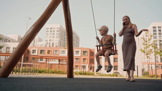 Woman talks on her phone while young boy swings at playground. scene takes place outdoors with modern apartment buildings in background featuring soft daylight and casual attire for both