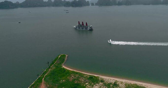 Aerial drone view showing a water plane speeding past a traditional cruise ship, with a sandy beach point in the foreground and limestone karsts rising across Ha Long Bay, Vietnam.