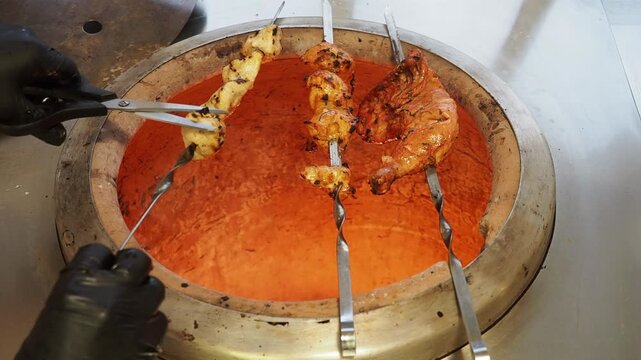 Chef rotates marinated chicken skewers inside a clay tandoor, cooking tikka and tandoori chicken over intense radiant heat in a professional Indian kitchen, close up static shot.