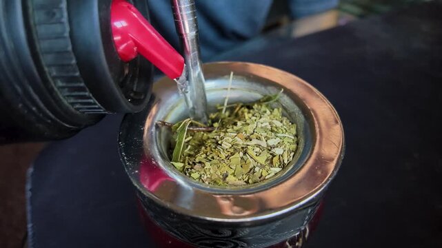 Yerba mate fills a traditional gourd as hot water is poured beside a filtered metal bombilla, showing traditional infusion preparation and beverage culture, static close-up.