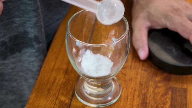 Hand adding creatine powder to an empty glass, then pouring in water and stirring it together as part of a daily 5 gram supplement routine, static shot