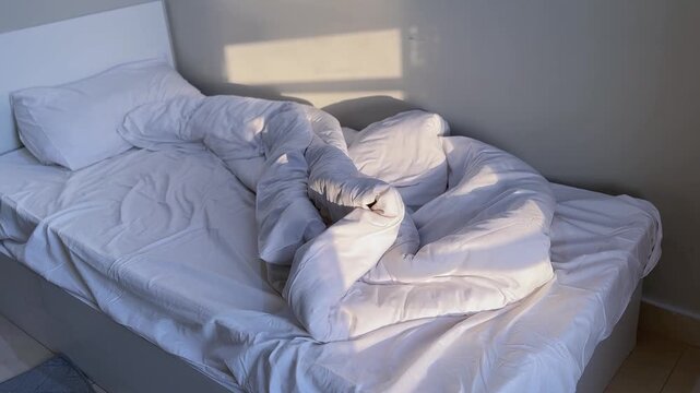 An unmade single bed with messy white sheets and a duvet sits in a minimal bedroom. Warm morning sunlight streams through a window, casting bright shadows on the wall during a peaceful daily routine.