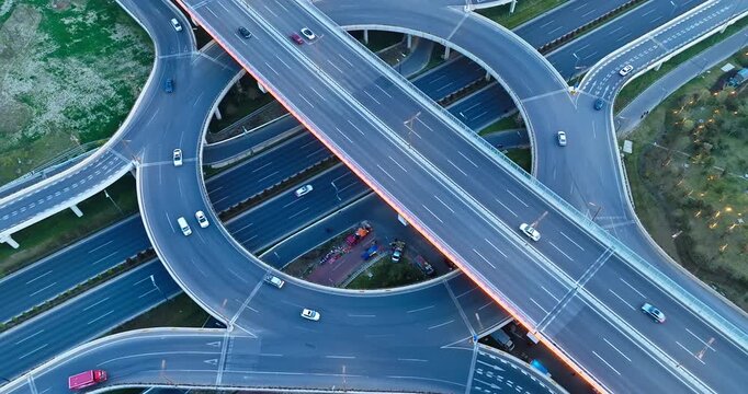 Aerial view of a multi-level highway interchange with curving flyover ramps, lane markings, passing vehicles, and bright natural evening light.