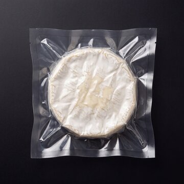 A round of creamy brie cheese, sealed in vacuum-packed plastic, showcases its delicate white rind against a dark background.