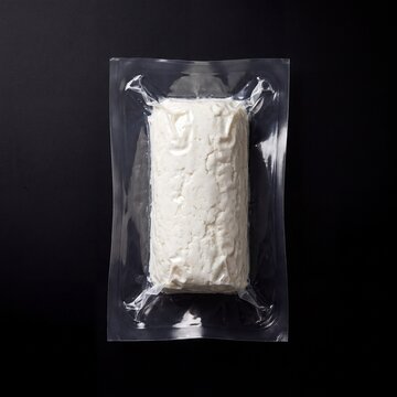 A rectangular block of fresh white cheese, possibly goat cheese, is sealed in clear vacuum-packed plastic packaging, highlighting its texture and form against a stark dark background.