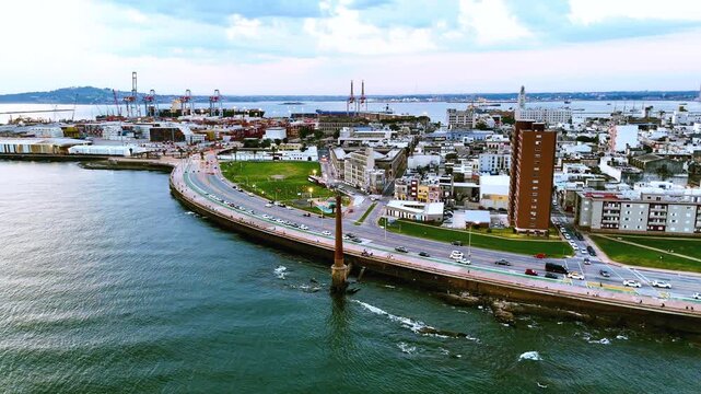 Drone view of Montevideo&rsquo;s waterfront showing the coastal road, urban buildings and the commercial Port of Montevideo with container terminals and harbor cranes at dusk.