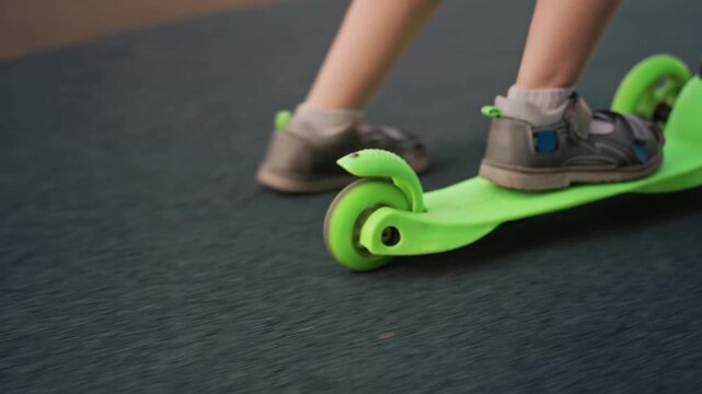 Young child rides bright green scooter across various surfaces including asphalt and wood. focus remains on lower legs and feet in motion as they propel or balance on three wheeled vehicle