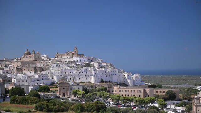 Centro Storico di Ostuni rises as a dense white hilltown above modern buildings and parked cars with the Adriatic on the horizon.