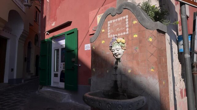 Decorative wall fountain with a smiling face spout stands in Piazza dei Martiri as sunlight hits a nearby green doorway in Procida.