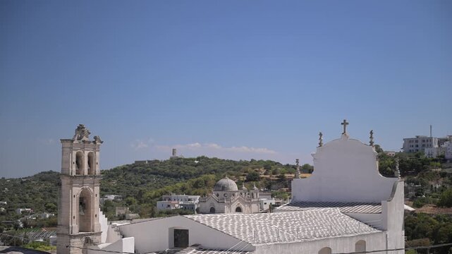 Static view of Ostuni&rsquo;s whitewashed church roofs and bell tower against tree-covered hills on a clear summer day.