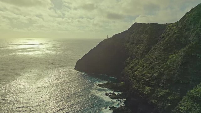 Sweeping drone aerial footage glides over choppy Pacific waters toward Makapuu Point&rsquo;s dramatic volcanic cliffs and iconic lighthouse on Oahu, Hawaii, bathed in warm golden hour sunlight.