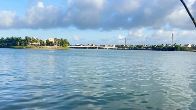 Eye-level wide shot gliding smoothly across Thu Bon River, featuring tranquil water, wooden fishing pole, lush green banks, Hoi An buildings, and bright morning sunlight under dramatic clouds.