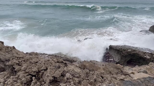 Powerful ocean waves crash against rugged black lava rocks on Oahu, Hawaii, sending white sea foam swirling through coastal tide pools in a dramatic low-angle shot under overcast skies.