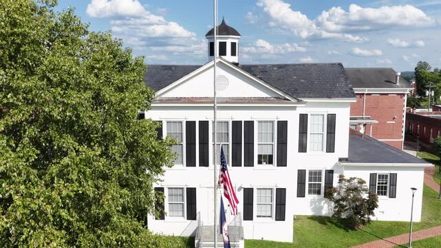 American flag flies before white historic courthouse building in Amherst, wooden cupola rises above pitched roof beside brick annex, tall shade tree lines front lawn area. Aerial drone view.