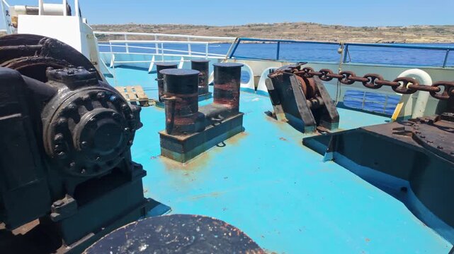 Gozo, Malta ferry deck with anchor chains, ropes, winch and mooring bollards on a sunny day in the Mediterranean harbor, with sea views and coastal transport context.