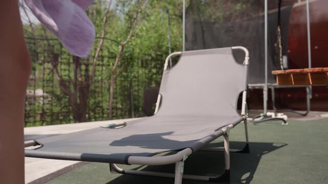 Person steps out of gray outdoor lounge chair with metal frame holding purple towel. scene is set in outdoor backyard or patio area with green foliage and fencing visible through background