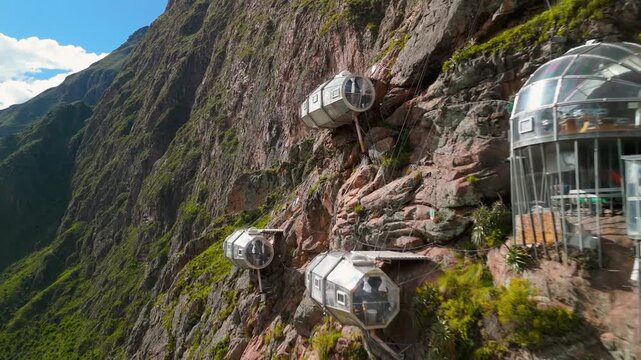 An aerial drone flies along a steep mountain cliff face, approaching the famous Sky Lodge Adventure Suites, a unique hotel of transparent glass pods offering extreme lodging in the Sacred Valley, Peru