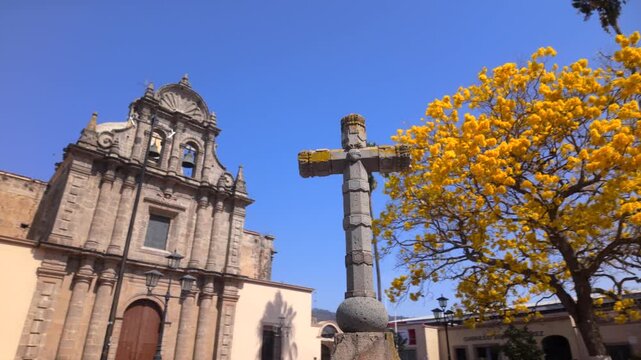 Stone church facade and plaza cross in Ciudad Guzman, Jalisco, Mexico, with golden tree and blue sky for travel, architecture, and city footage.