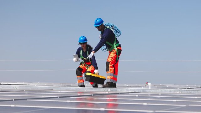 Two solar technicians working on rooftop photovoltaic installation, one drilling panel mounts while colleague checks system data on a tablet.