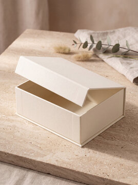 Elegant open ivory rigid gift box with a partially lifted lid, styled on a textured stone surface with eucalyptus and dried botanicals in warm natural light.