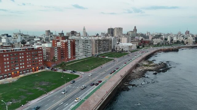 Drone view along Montevideo&rsquo;s waterfront boulevard at dusk, showing coastal traffic, promenade, urban buildings, green spaces and the Port of Montevideo in the distance.