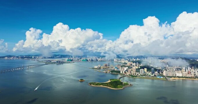 Aerial view of a coastal city with bridges and a small green island under vast cumulus clouds during bright sunny weather.