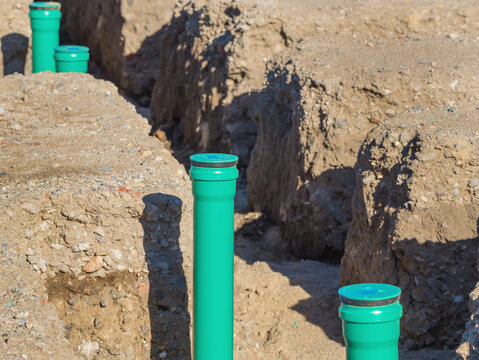 Civil engineering site featuring an underground utility trench for city canalization. Vertical green plastic sewer pipes installed in an excavated dirt ditch for municipal wastewater management.