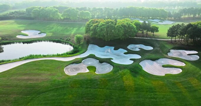 Aerial view of a lush green golf course featuring white sand bunkers, curved pathways, and pond under bright morning sunlight.