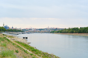 Obraz premium Wide scenic landscape of the calm blue Danube river flowing through Budapest towards Margaret Bridge with distant historic architecture and green embankments under a light cloudy sky. 