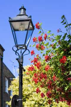 Traditional black street lamp post decorated with bright red hanging geranium flowers against a blue sky