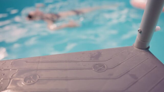 Swimmers move through clear blue water as pool entry step with metal pole and embossed symbols remains in focus. Water ripples naturally suggesting active swimming motion throughout scene