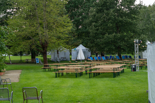 A wide view shows a beer garden layout with wooden benches, deck chairs, a closed umbrella and white event tents beneath trees.