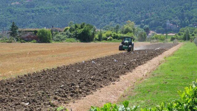 Tractor plowing a harvested wheat field in rural Liguria, Italy, turning dry stubble into dark soil with mountains and countryside in the background. Agriculture concept, agribusiness.