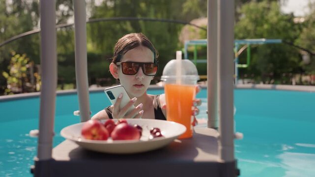 Young woman relaxes in pool taking selfies with her smartphone while surrounded by fresh fruit and orange drink. Woman adjusts phone for better angles as woman enjoys leisure time outdoors