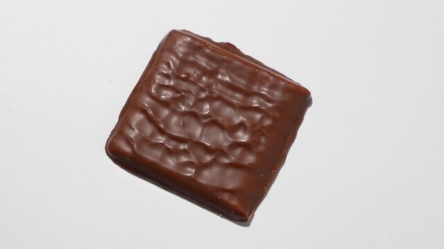A single square chocolate-covered cracker lies flat on a white surface and slowly rotates, showing its glossy textured coating from above.