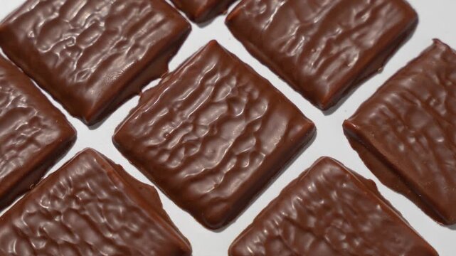 Chocolate-covered crackers are laid out in a grid on a white surface and rotate slowly together, filmed in extreme macro close-up detail.
