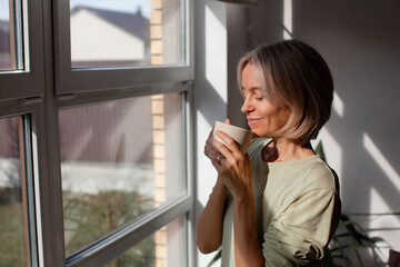 Obraz premium Middle aged woman cradles cup beside bright window, showing relaxed smile and closed eyes. Female adult takes peaceful morning pause indoors, warming hands around drink, holding mug in front of face.