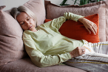 Middle aged woman resting on sofa appears tired while holding orange pillow. Female adult reclines...