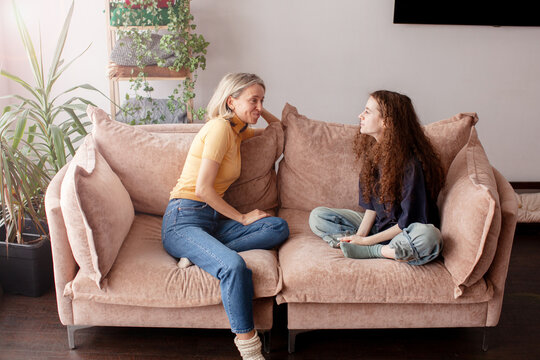 Smiling middle aged woman talks with teenage girl seated cross legged on sofa during family conversation. Woman maintains eye contact while girl responds attentively in bright living room.