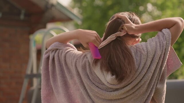 Young woman with long brown hair ties her ponytail outdoors wearing light purple bathrobe. Woman uses both hands to gather and secure her wet hair against backdrop of green foliage and part