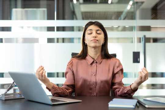 Young businesswoman meditating at her office desk with eyes closed, finding calm and stress relief amidst work, practicing mindfulness for mental wellness and focus