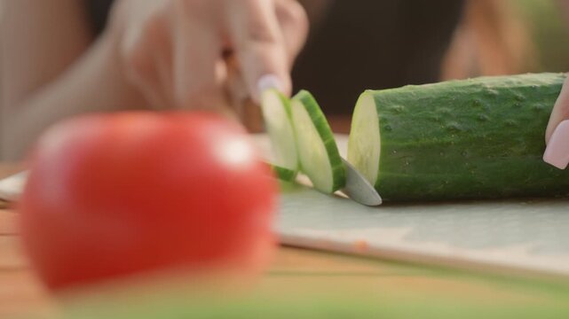 Woman slices fresh cucumber on cutting board with soft natural light while ripe red tomato is visible in foreground. Close up view emphasizes texture detail and precise knife work