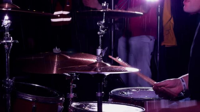 Drummer striking cymbal and drums amid water spray under purple concert lights