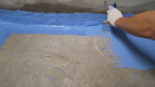 Worker in glove applying blue waterproofing membrane to concrete floor and wall joint with paintbrush during renovation