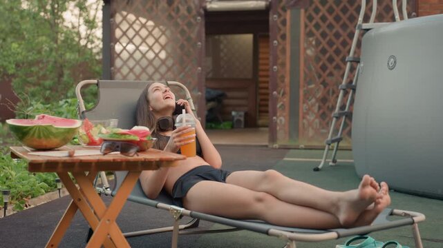 Young woman relaxes on lawn chair outdoors holding orange drink and talking. wooden table beside her holds watermelon slices sunglasses food items and drinks in clear containers. background